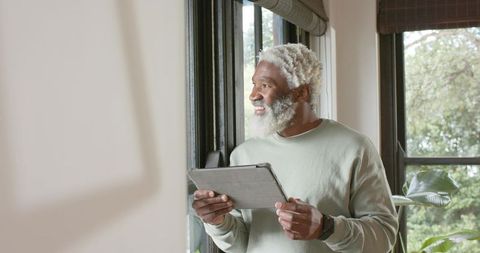 Elderly Man By Window Holding Tablet Near Indoor Plant