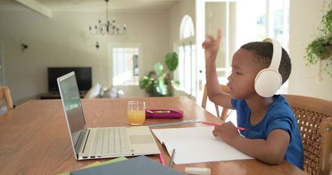Young african american boy raising hand during remote learning with headphones and laptop