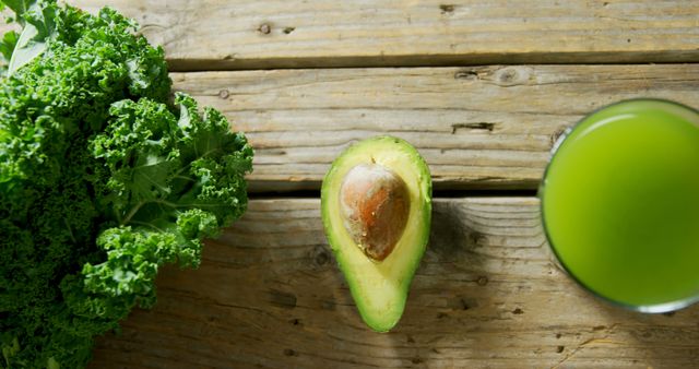 A halved avocado and a bunch of kale are placed next to a glass of green juice on a wooden surface, with copy space. These fresh ingredients are often associated with healthy eating and detox diets.