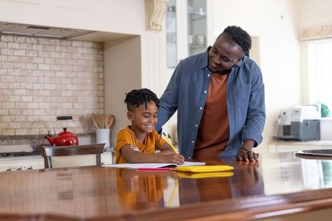 African american father helping son with homework at kitchen island, smiling and writing