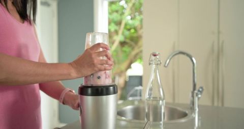 Woman blending berry smoothie at kitchen counter amid sunlit window and stainless faucet