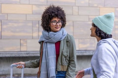 Friends greeting and chatting on urban plaza with rolling suitcase, scarves and beanie
