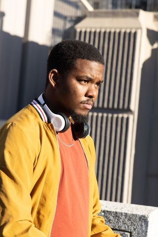 Sunlit african american man standing on rooftop wearing mustard jacket and headphones