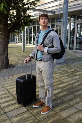 Young traveler waiting with suitcase and backpack outside modern transport hub