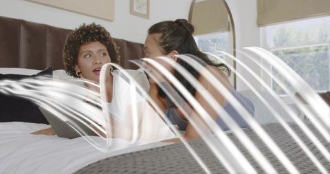 Chatting women reclining on bed holding tablet and laptop sharing bedroom conversation