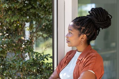 African woman gazing out window in rust shirt and gold hoop earrings, indoor natural light