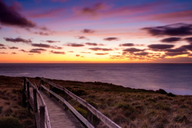 Image shows a peaceful twilight scene with a wooden walkway leading towards the ocean. The vibrant sunset colors fill the sky, reflecting over the water to create a serene and tranquil atmosphere. Ideal for travel brochures, nature documentaries, scenic calendars, or meditation backgrounds.