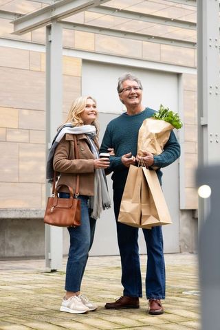 Senior couple linking arms carrying groceries and coffee on urban weekend stroll