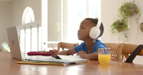 African american boy studying remotely wearing headphones at home with laptop and notebook