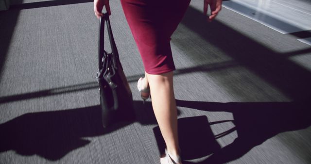 Businesswoman in a maroon skirt walking on an urban walkway, holding a black handbag. Ideal for illustrating business travel, professional lifestyle, urban commuting, and corporate environments.