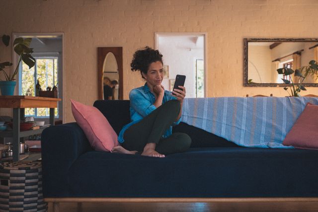 Biracial woman sitting comfortably on a sofa, using a smartphone in a cozy living room. Ideal for depicting home isolation, quarantine lifestyle, modern communication, and casual relaxation. Suitable for articles on remote work, staying connected, or home decor.