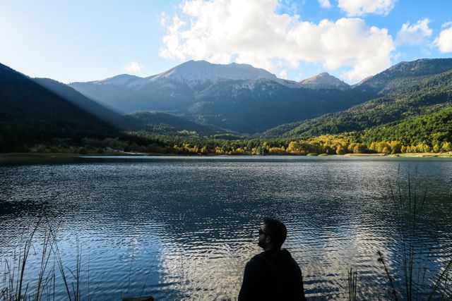 Person admiring calm lake with mountains in background under sunlit sky. Useful for themes of tranquility, nature appreciation, outdoor activities, and introspection. Ideal for travel blogs, adventure promotions, nature conservation campaigns, wellness programs, and outdoor lifestyle magazines.