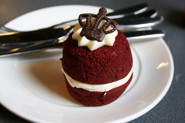 This elegant red velvet cupcake is topped with cream cheese frosting and a decorative chocolate crown, presented on a white plate. Ideal for bakery advertisements, gourmet recipe blogs, and celebration event promotions.