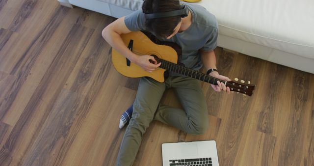 Young man sitting on floor playing guitar while wearing headphones and using laptop in cozy home environment. Great for topics related to online music lessons, remote learning, personal hobbies, and home activities.