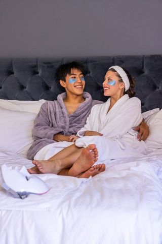 Couple relaxing on bed wearing under-eye patches and led face mask in plush bathrobes