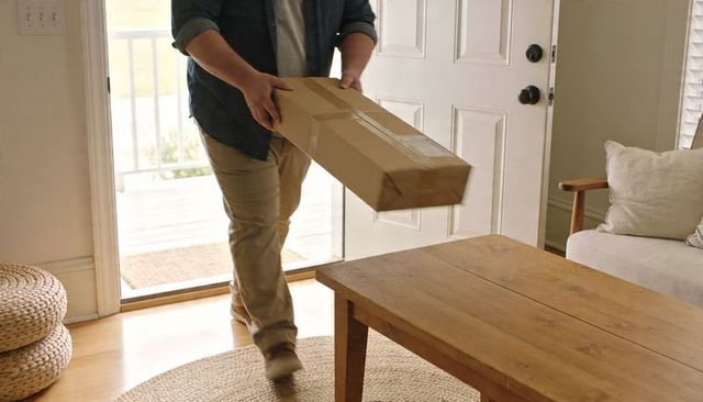 Man carrying long shipping box into cozy living room, delivering parcel at front doorway