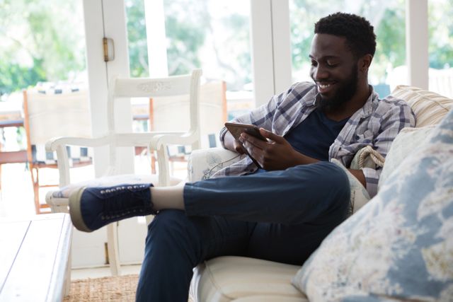 Man sitting on a couch in a bright living room, using a digital tablet. He is smiling and appears relaxed, enjoying his time at home. This image can be used for lifestyle blogs, technology advertisements, home decor websites, or articles about modern living and relaxation.