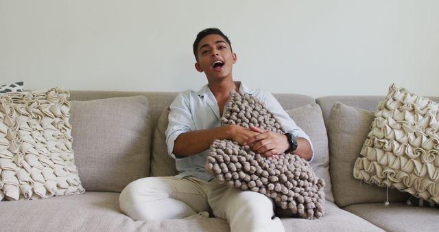 A young man is sitting on a beige couch in a relaxed manner, holding a textured cushion and singing. The setting appears to be casual and comfortable, making it suitable for lifestyle and leisure themes. This can be used for promotions related to home comfort, relaxation tips, casual living, or joyful moments at home.