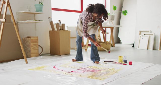 Artist is creating a large painting on canvas laid on the floor of a bright, spacious studio. Surrounded by various art supplies, the artist appears focused and deeply engaged in their work. Perfect for illustrating creativity, artistic process, modern art studios, or DIY project inspiration.