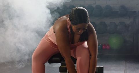 Focused african american woman resting on bench after intense workout in moody gym