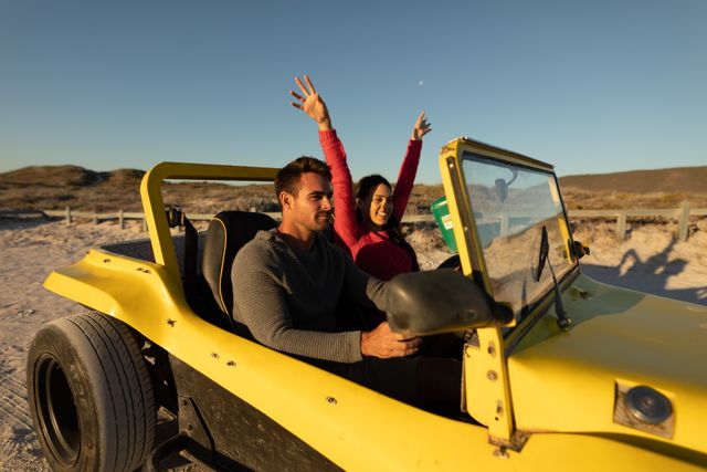 Happy caucasian couple driving in beach buggy on sunny beach, smiling and raising arms. beach stop off on romantic summer holiday road trip.