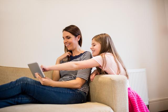 Mother and daughter sitting on sofa spending quality time together using tablet. Ideal for family, technology, and lifestyle content showcasing the use of digital devices in a home environment. It can be used in articles about parenting, family activities, or modern living.