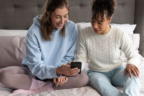 Multiracial friends sharing smartphone on bed wearing cozy loungewear in pastel bedroom