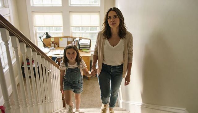Mother guiding daughter hand-in-hand up carpeted stairs with home study in background