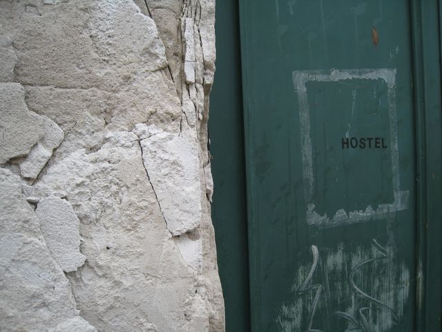 Close-up of an old, weathered door with 'Hostel' marked on it next to a textured stone wall. Image captures the rustic and distressed look of the materials used. Perfect for use in projects related to urban exploration, historic buildings, and vintage or rustic aesthetic themes.