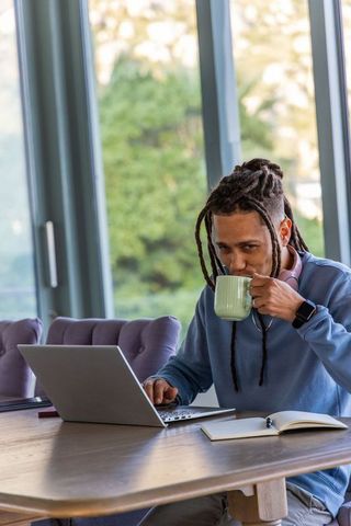 African american man sipping coffee while working on laptop in sunlit home office