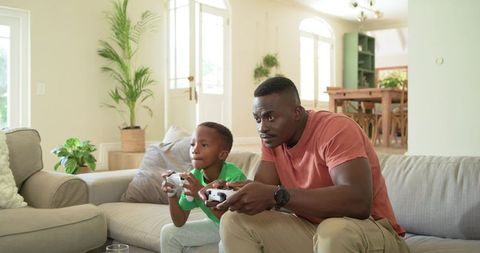 African american dad and son playing video games on couch enjoying focused gaming