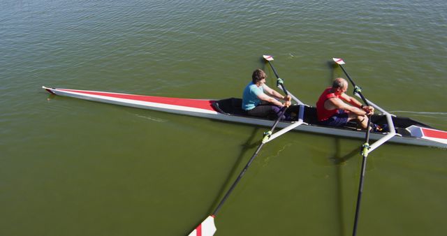 Two individuals are rowing a narrow boat together on a calm lake, showcasing teamwork and coordination in an outdoor setting. Ideal for use in articles or advertisements about fitness, outdoor sports, teamwork, and recreational activities. It can also be used for promoting rowing clubs or for educational content about water sports.