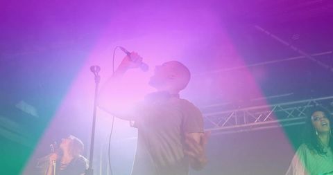 Bald male vocalist singing with headphones holding microphone under magenta purple green lights