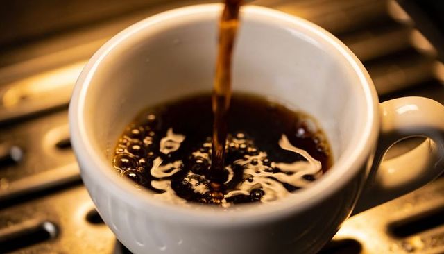 Pouring dark coffee into white ceramic cup creating crema, foam and warm golden ripples