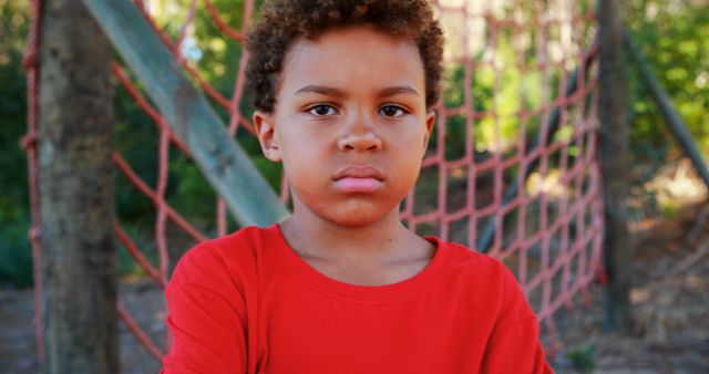 A young boy is standing at a playground with a red rope net behind him, wearing a red shirt and showing a serious expression. This image can be used in educational materials, children's activities promotion, child psychology articles, and advertisements for playgrounds or children's clothing.