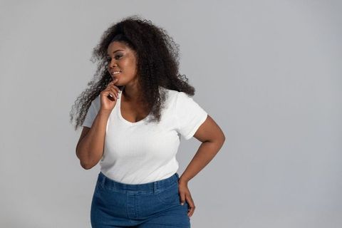 Confident african american woman posing in white ribbed top and denim jeans on neutral backdrop