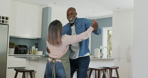 Mature diverse couple dancing and laughing in modern sunlit kitchen holding hands