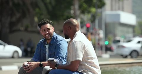 Male friends chatting and holding coffee while sitting on fountain edge in urban plaza