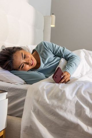 Asian woman relaxing in bed smiling at smartphone in cozy modern bedroom morning light