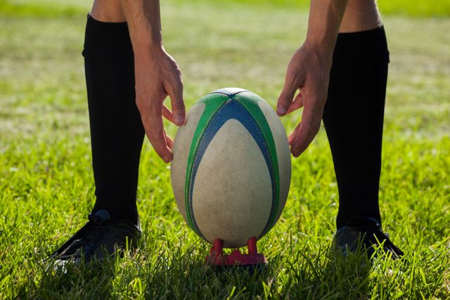 Rugby player placing ball on tee, preparing to kick for goal on grassy field. Ideal for sports articles, athletic training materials, and competitive sports promotions.