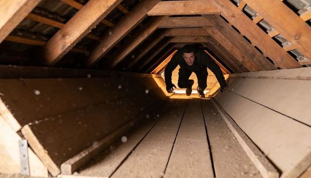 Crawling man inspecting narrow attic crawl space moving toward lit hatch and rafters