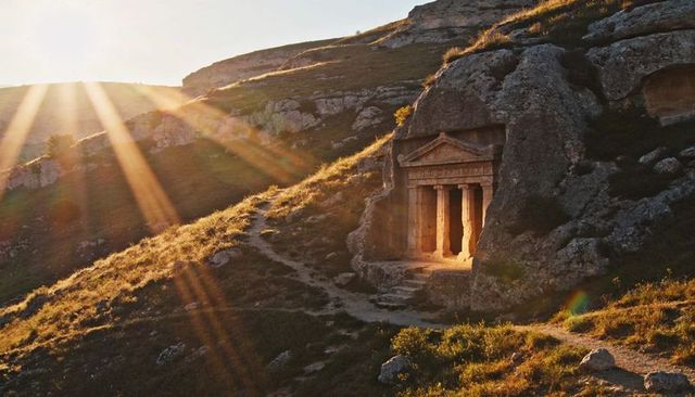 Rockcut temple facade glowing under sunrays on rugged hillside with ancient columns and path