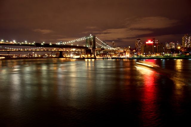 Stunning night view of the Brooklyn Bridge over the East River in New York City, with city lights reflecting on water. The long exposure gives a smooth, glossy effect to the water, adding a modern mystical touch. Ideal for use in travel promotions, architectural magazines, New York City guides, and urban landscape portfolios.