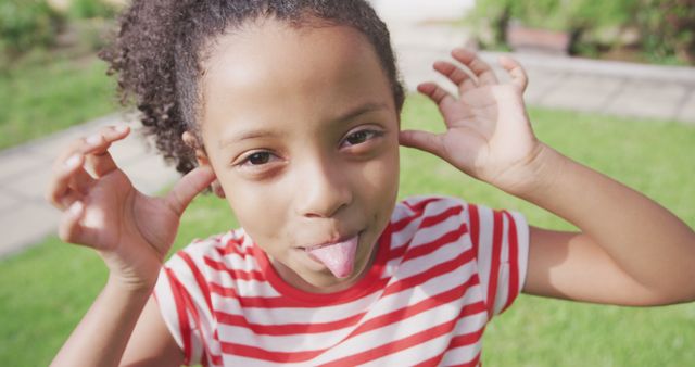 Young girl with curly hair making a silly face by sticking her tongue out and putting her fingers to her ears. She is wearing a red and white striped shirt and standing outside in a park. Image conveys childhood joy, playfulness and fun. Ideal for use in advertisements promoting children's products, playful activities, summer camps, or educational content related to child's development and well-being.
