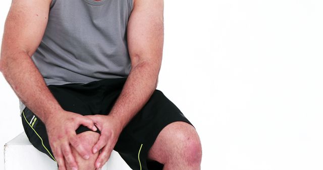 This stock photo depicts a man wearing a gray sleeveless shirt and black shorts, sitting and holding his knee in discomfort. The focus is on his hands and knee, highlighting issues related to knee pain, injury, and health. Useful for articles and materials discussing sports injuries, fitness health, or treatment for joint pain and arthritis.