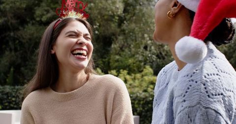 Friends laughing in sunlit backyard wearing merry christmas headband and santa hat