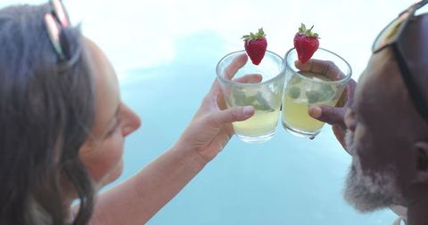 Clinking cocktails by poolside with strawberries and mint, diverse senior couple relaxing
