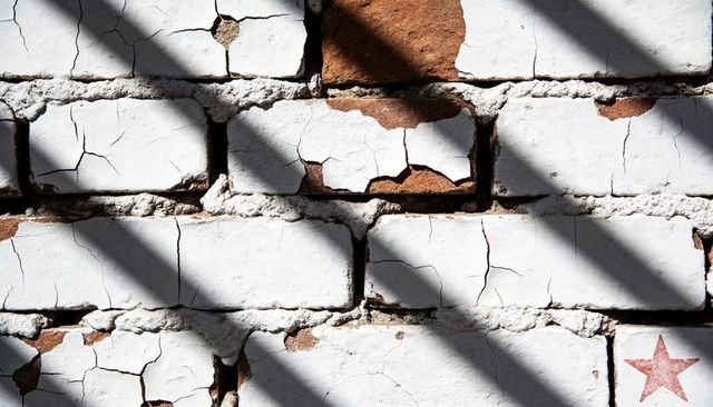Peeling white paint on red brick wall with diagonal shadows and faded red star