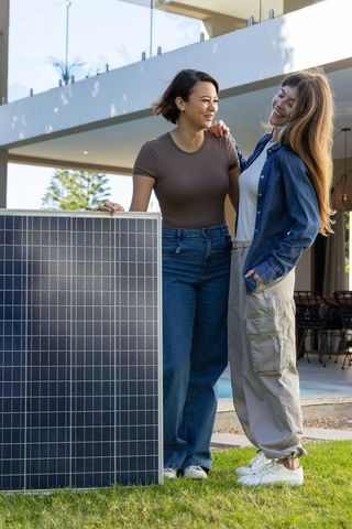 Female friends standing with solar panel in modern backyard promoting solar energy