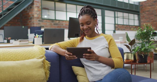 Young woman sitting on blue sofa in a modern office, holding and using a digital tablet while smiling. Her casual attire and relaxed posture suggest a comfortable and productive work environment. Suitable for themes such as modern workspaces, remote work, women in technology, and relaxation in office settings.